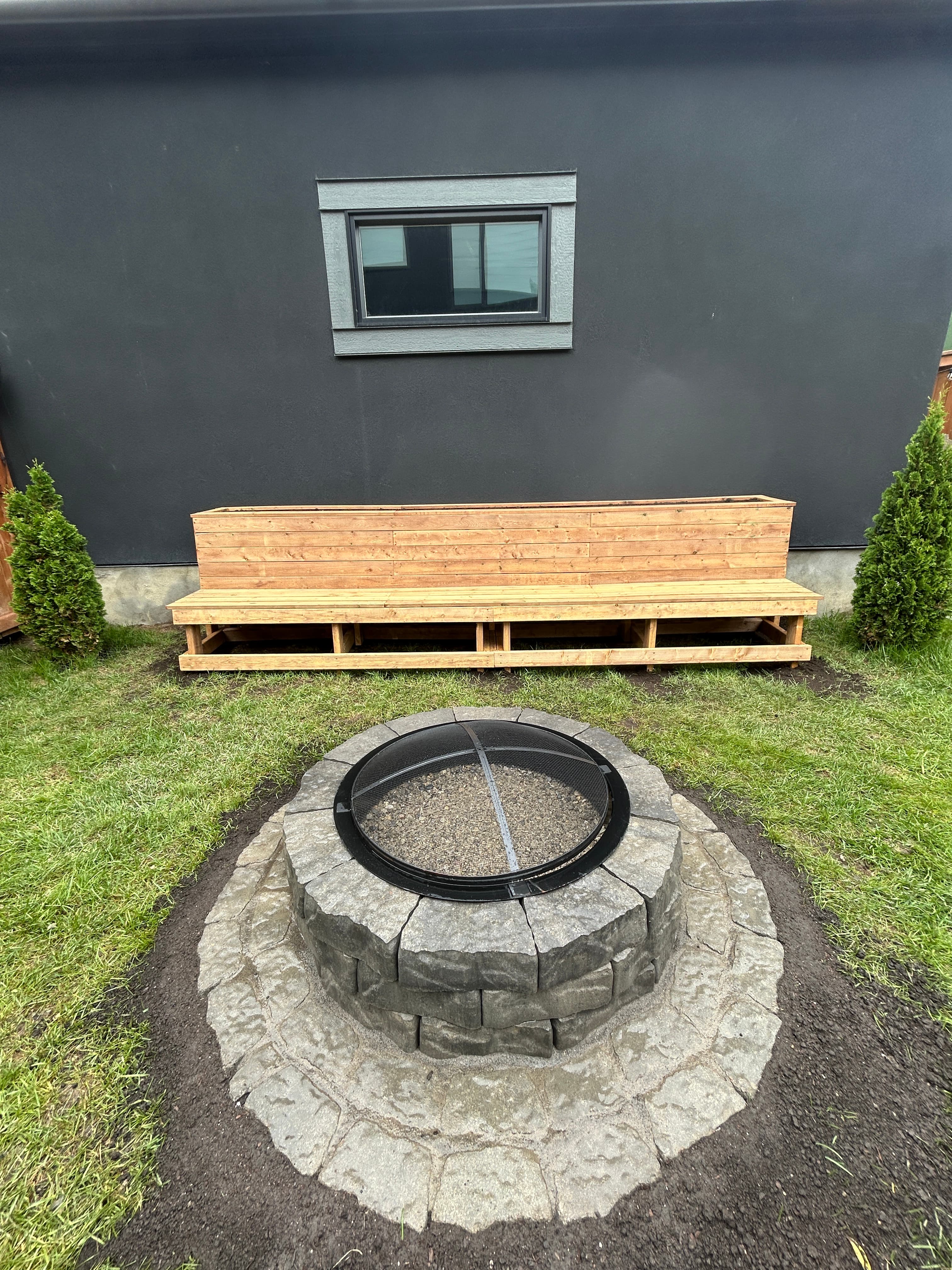 Stone fire pit with mesh cover beside a wooden bench against a dark grey wall.