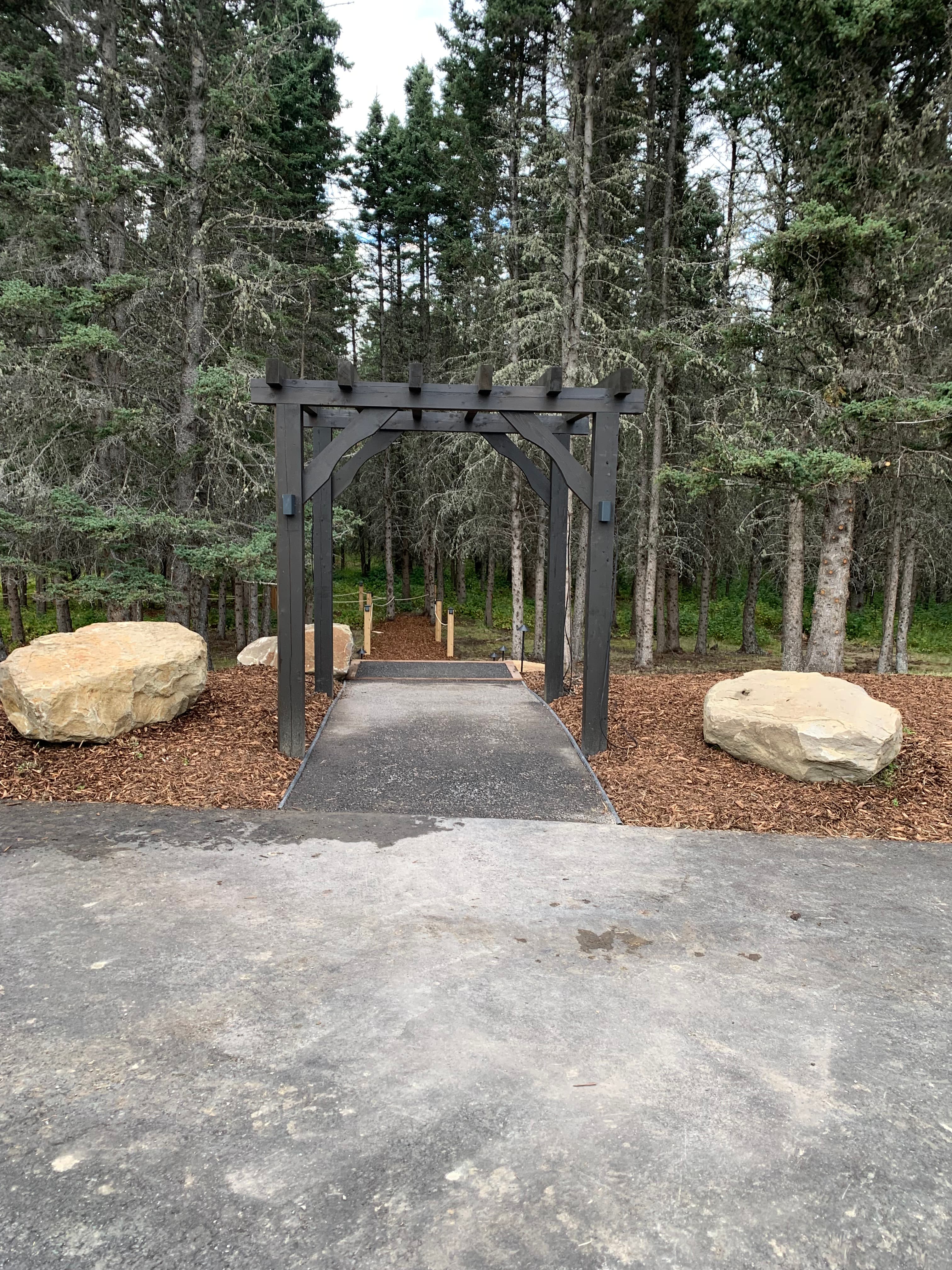 Dark wooden arbor over a paved path leading into a dense evergreen forest with rocks.