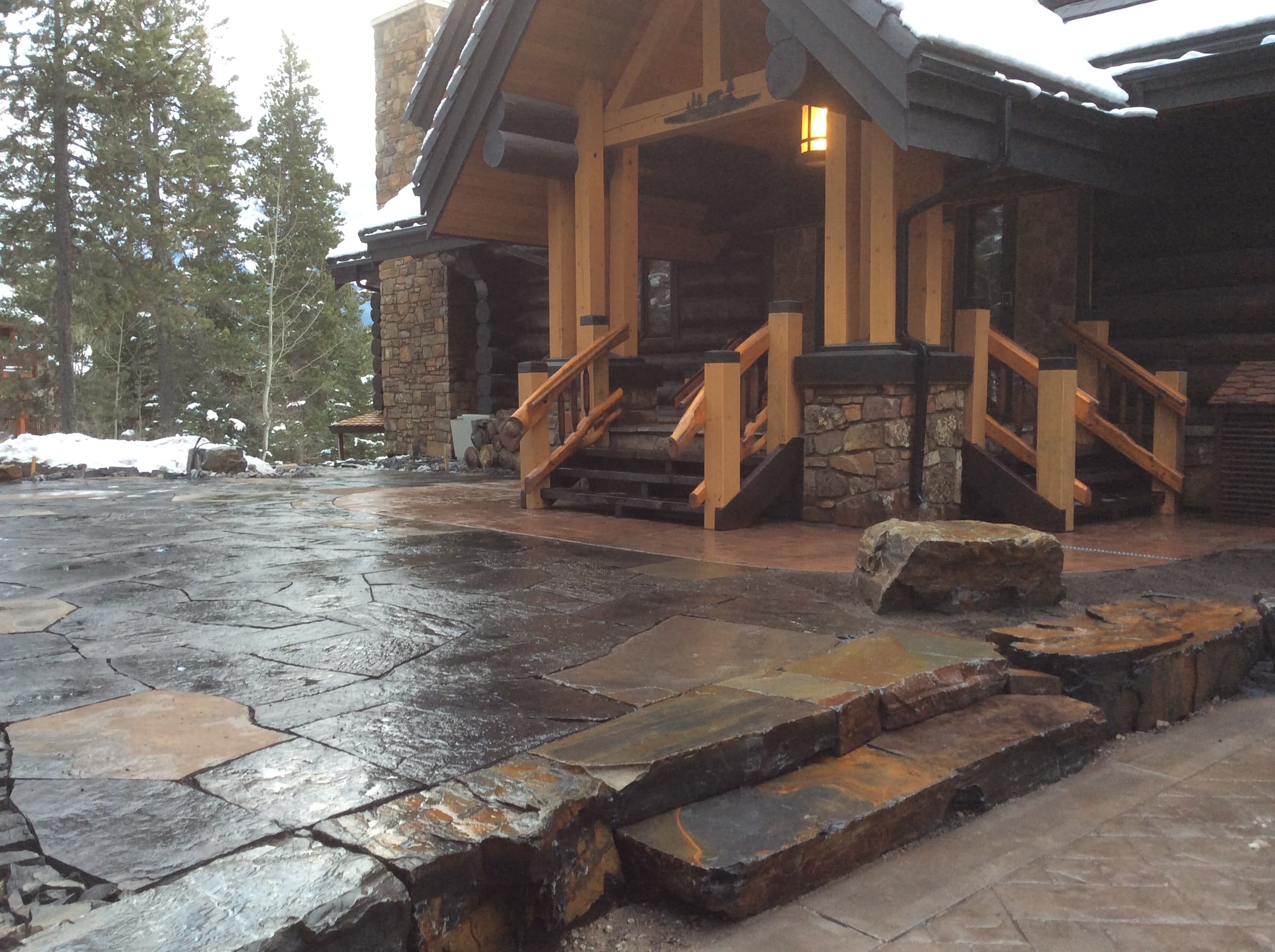 Rustic log cabin entrance with stone steps and a wet flagstone patio in winter.