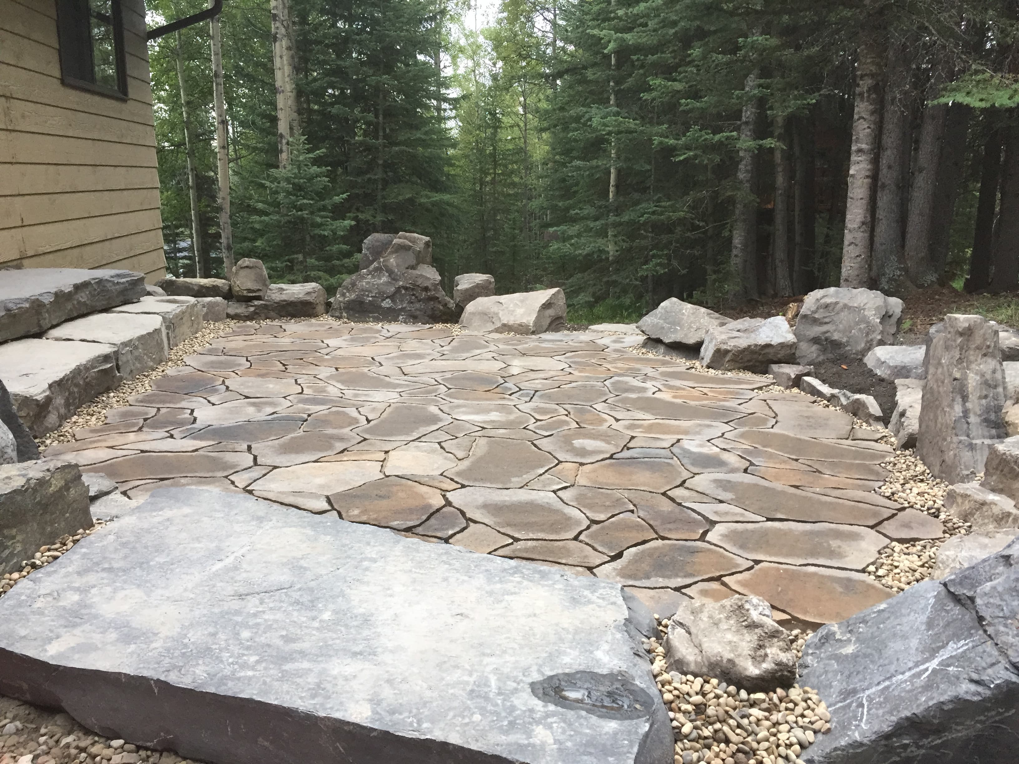 Rustic flagstone patio surrounded by large boulders and pebbles, set within a lush evergreen forest.