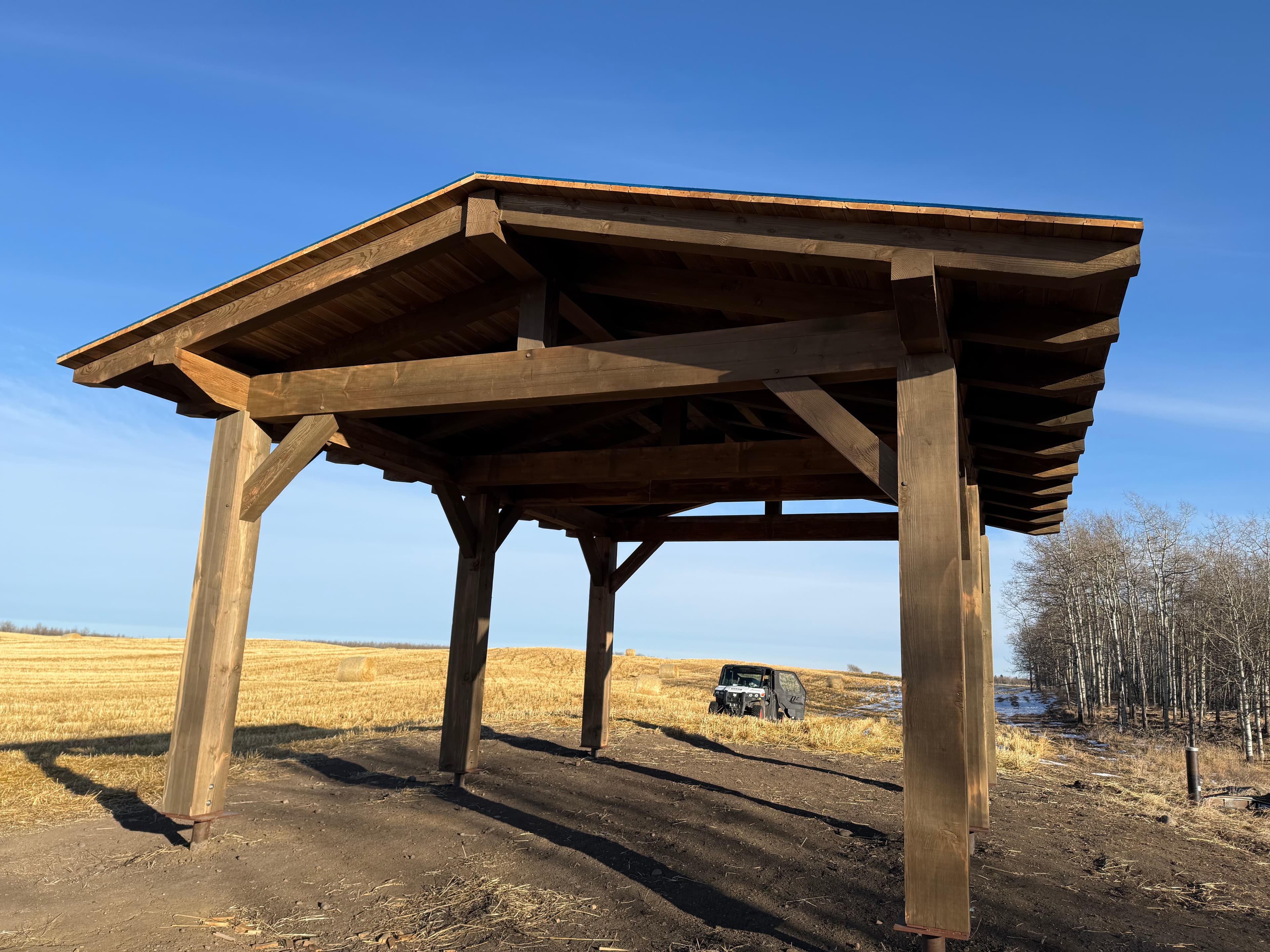 Large wooden timber frame pavilion in a harvested field under a clear blue sky.