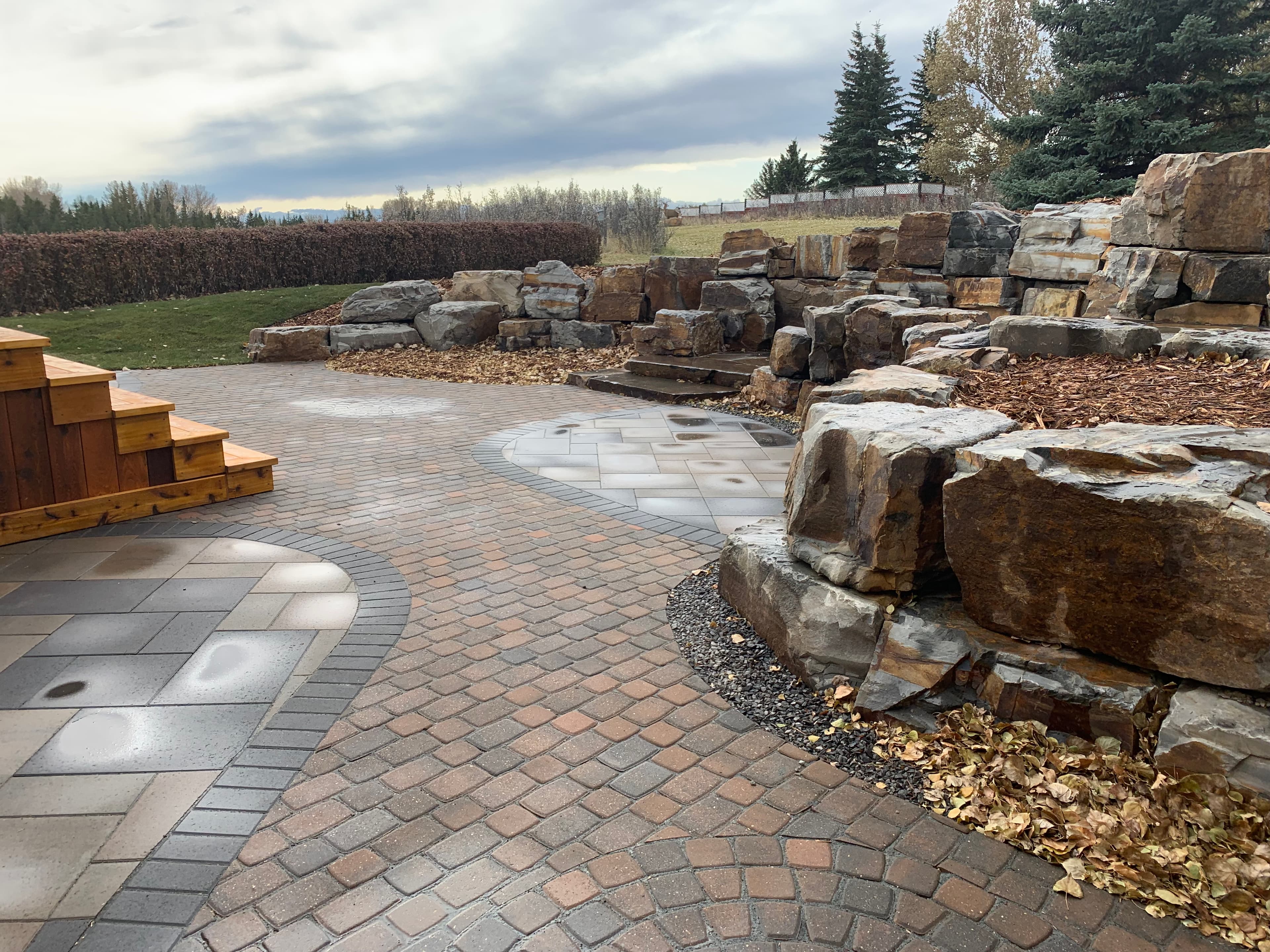 Multi-toned curved brick patio featuring a large stacked rock retaining wall and wooden steps.