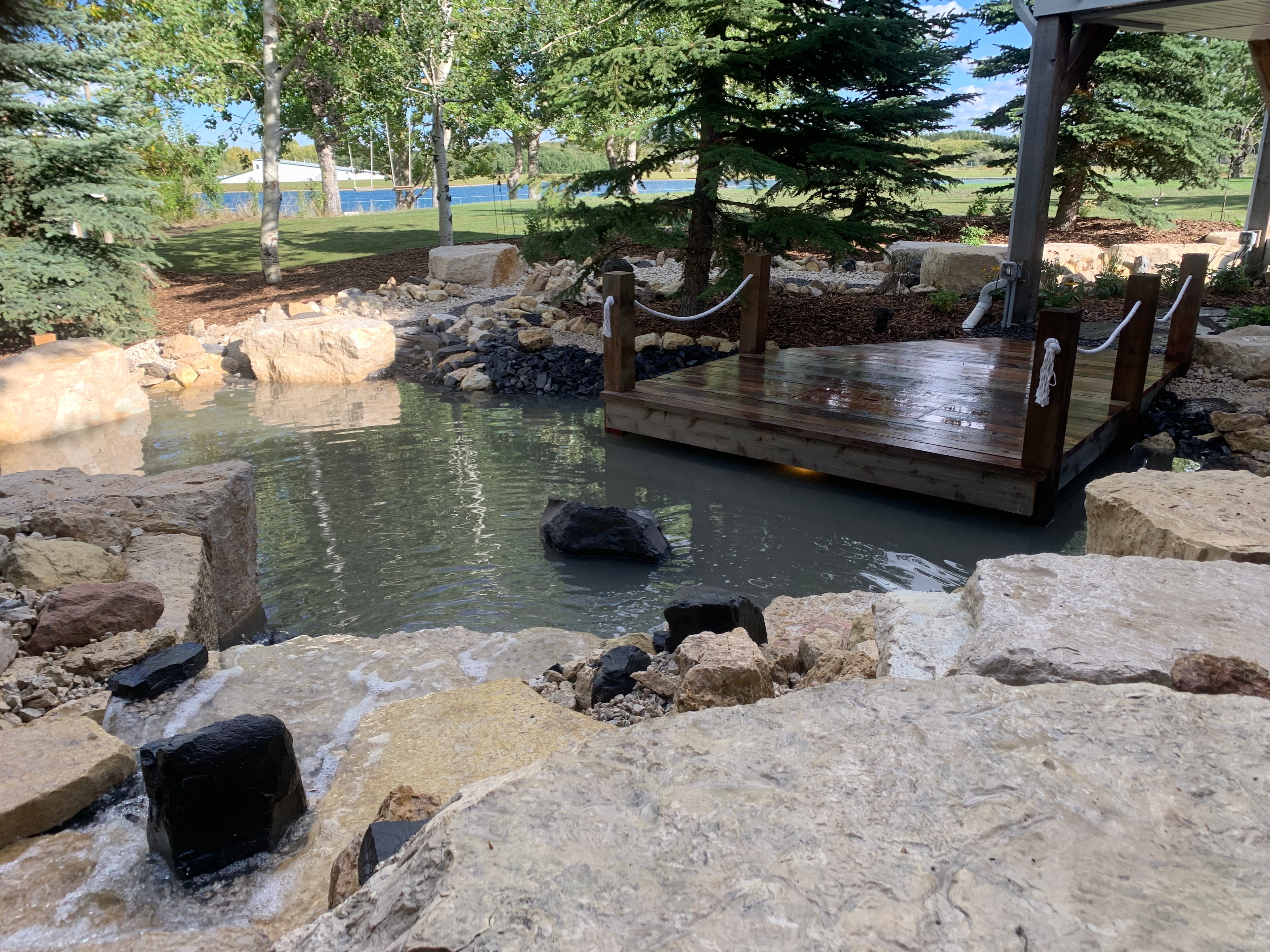 Wooden deck with rope railings over a rocky backyard pond surrounded by lush green trees.