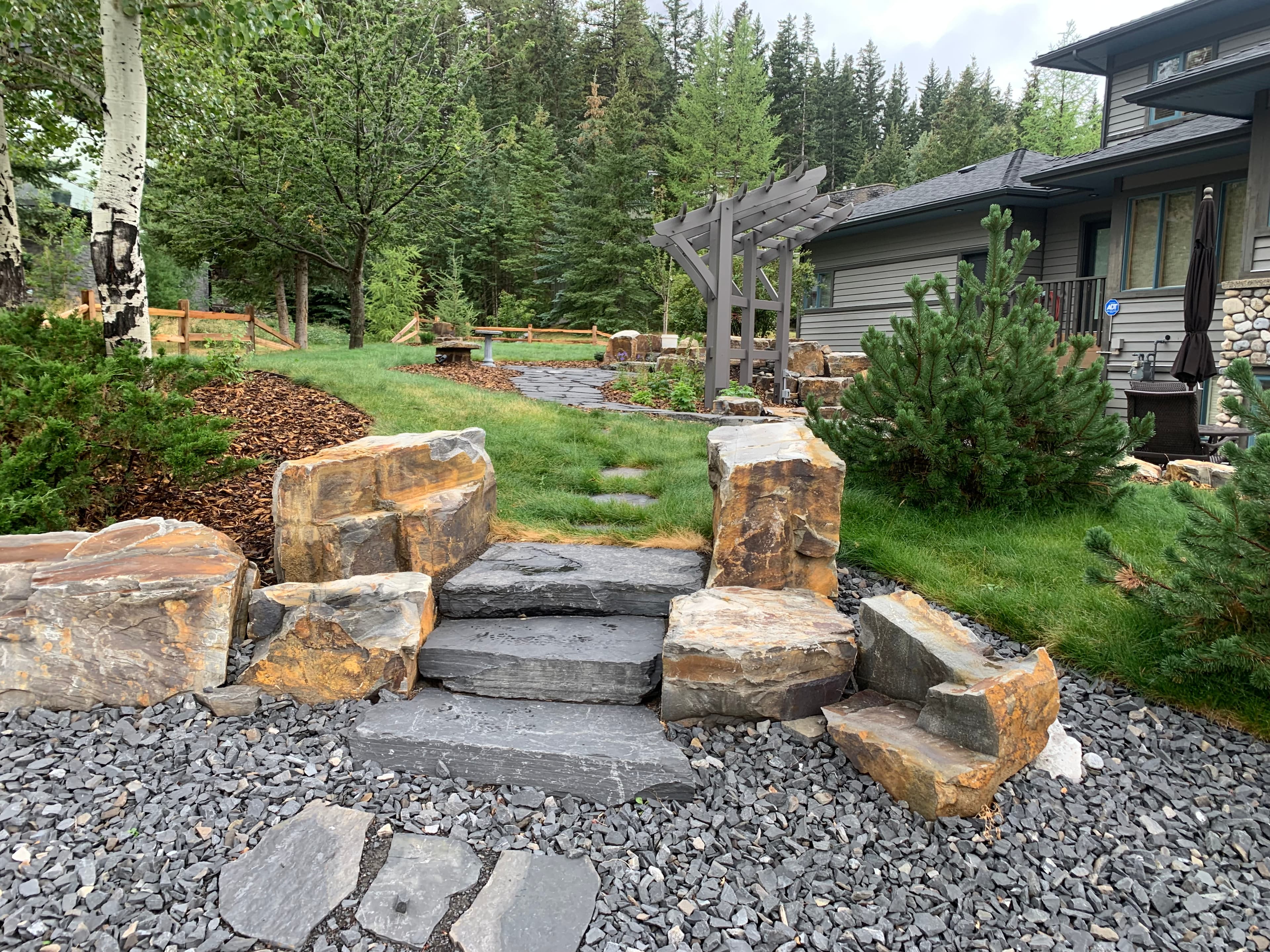 Slate stone steps and large rocks lead to a pergola and modern house in a forest.