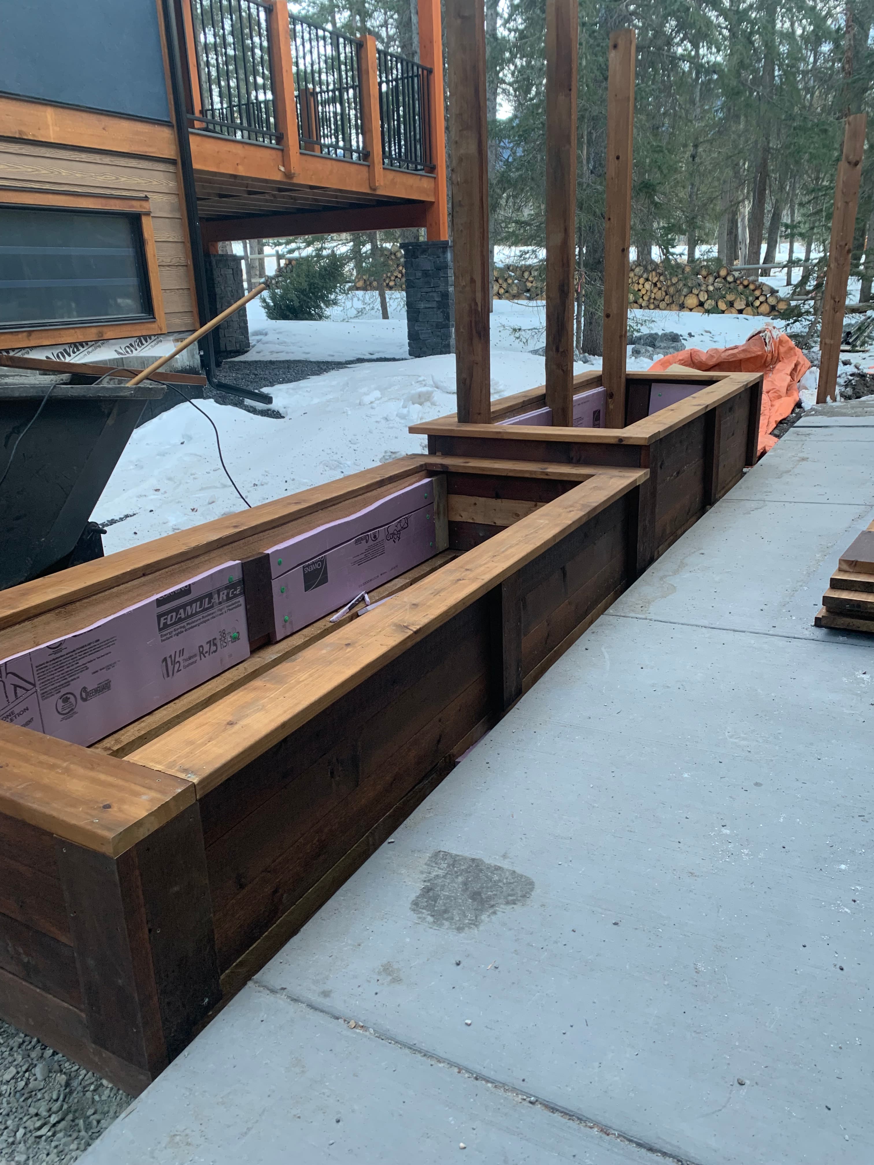 Long dark-stained wooden planter box lined with pink insulation boards on a concrete patio.