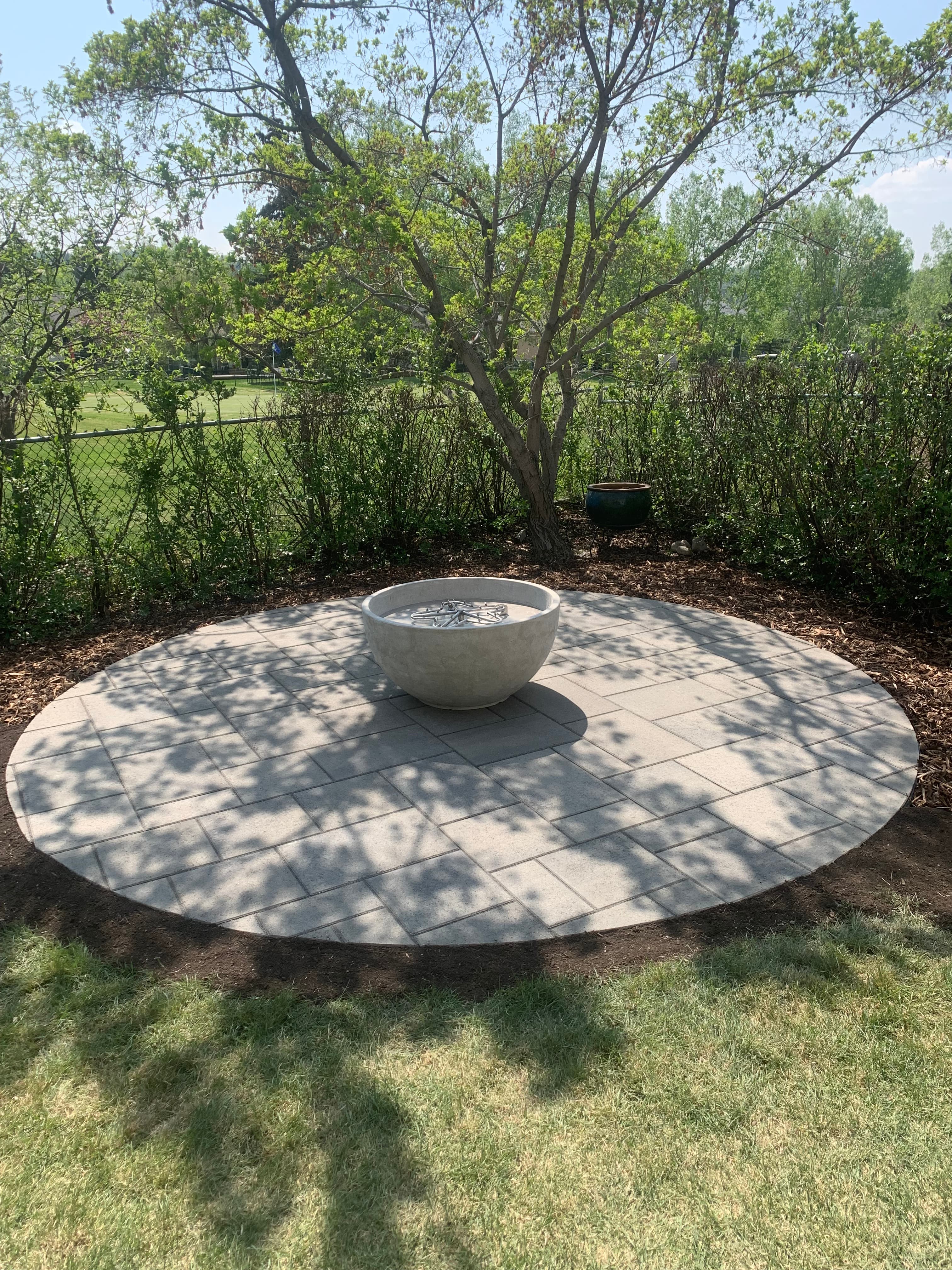 Circular stone patio with a concrete fire pit bowl, surrounded by lush green trees.