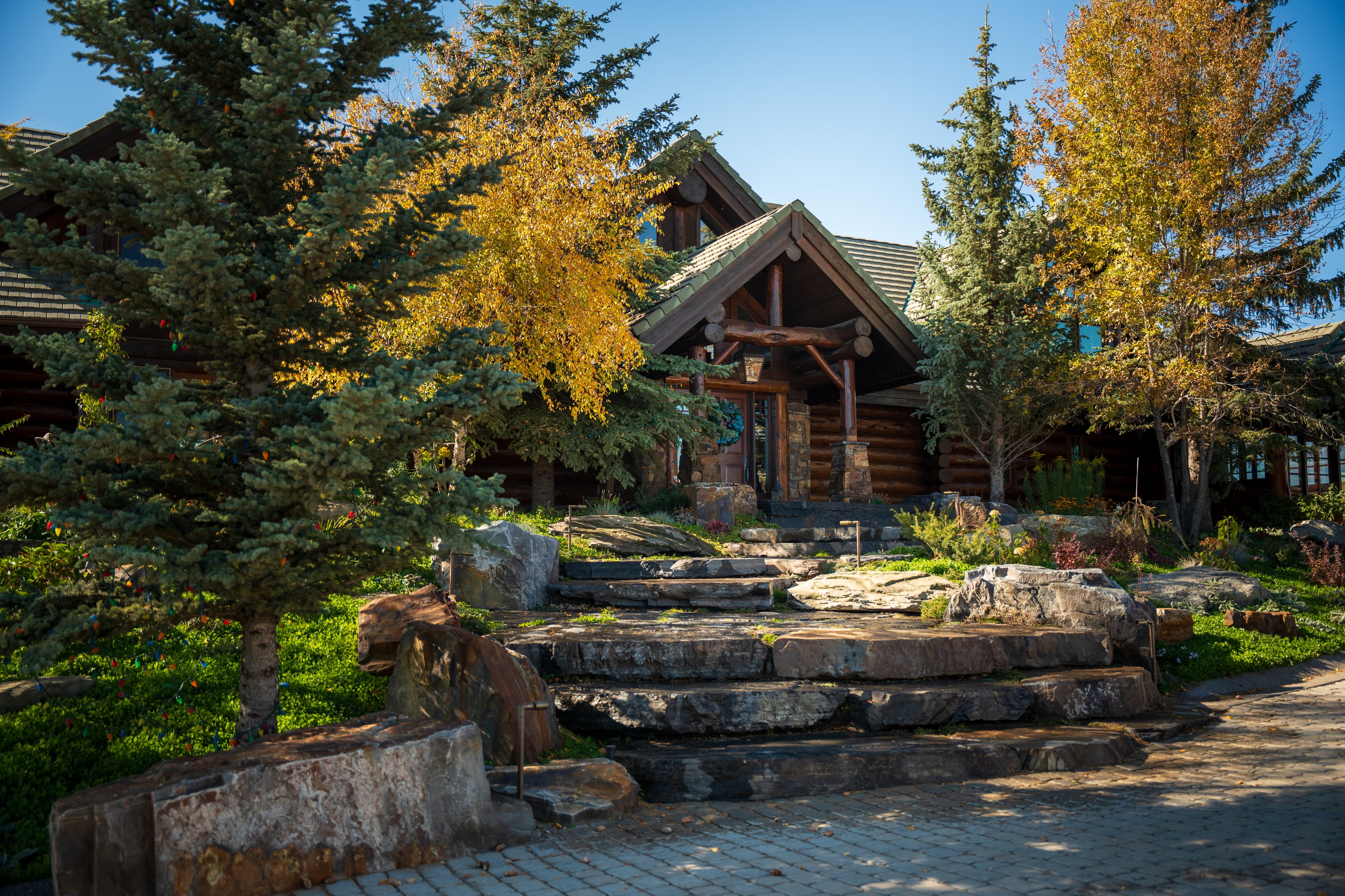 Rustic log cabin with stone steps and autumn trees under a bright blue sky.