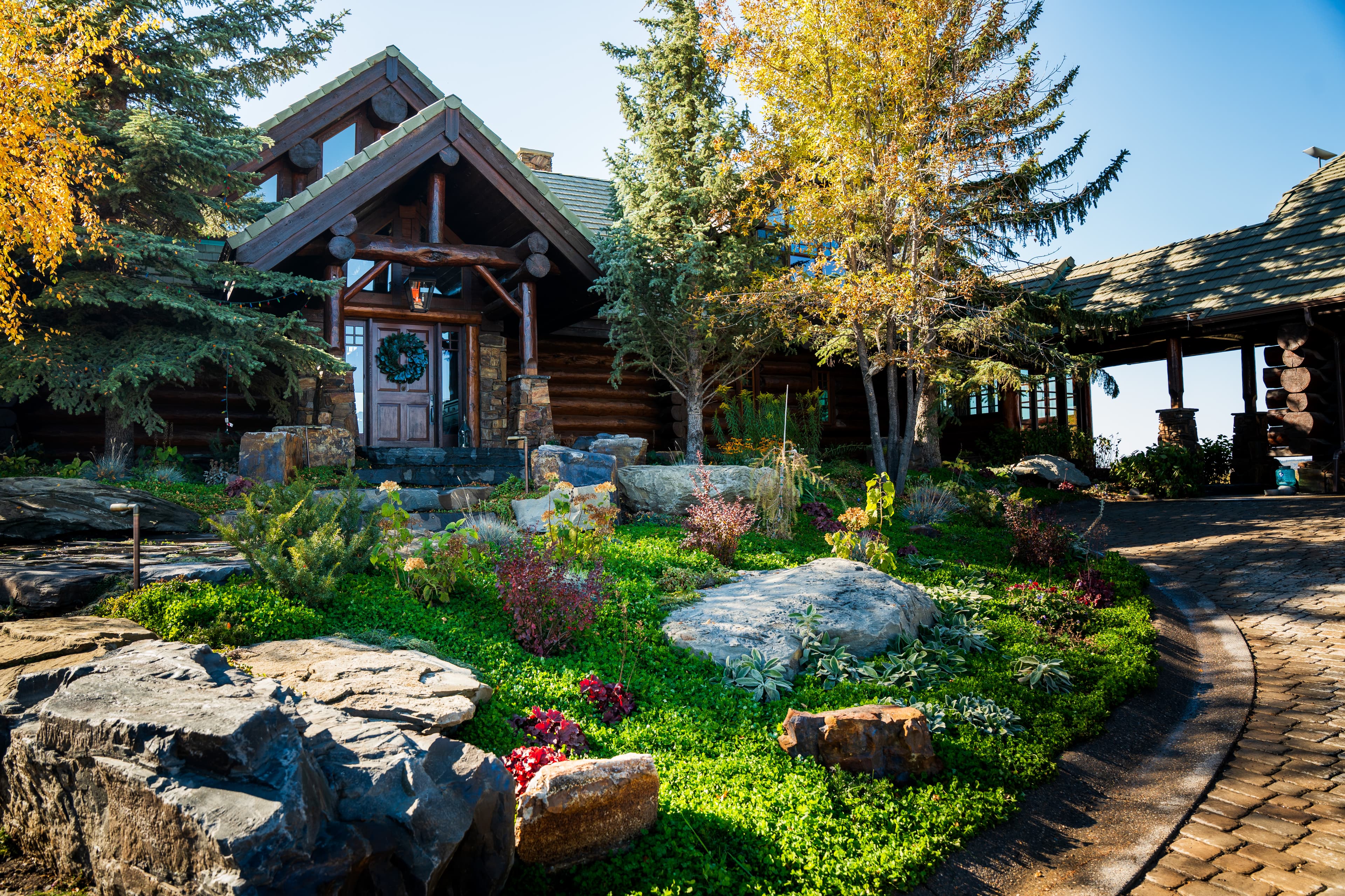 Rustic log cabin with stone landscaping, autumn trees, and a wreath on the front door.