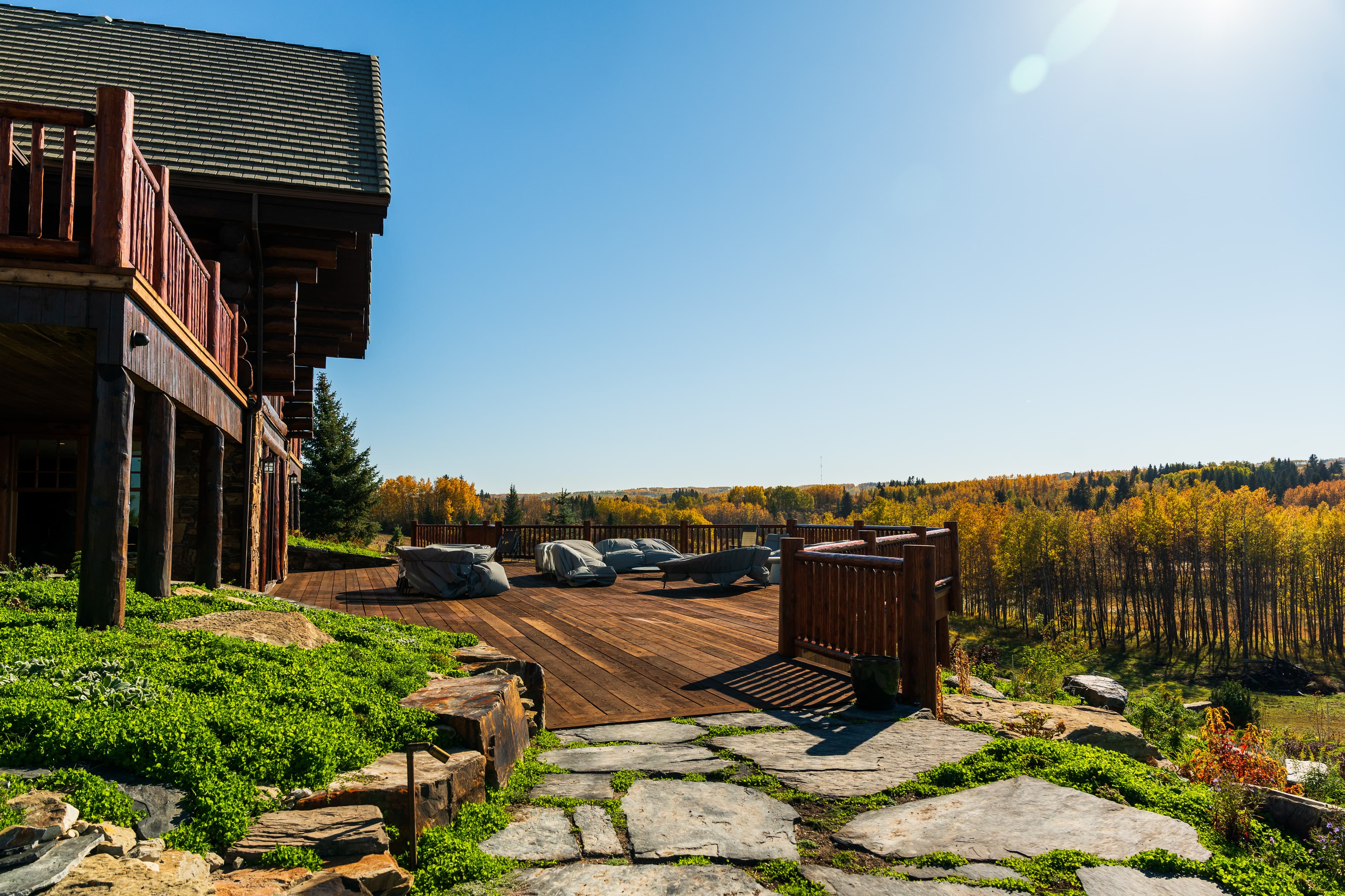 Large wooden deck with covered furniture overlooks a vibrant autumn forest under sunny skies.