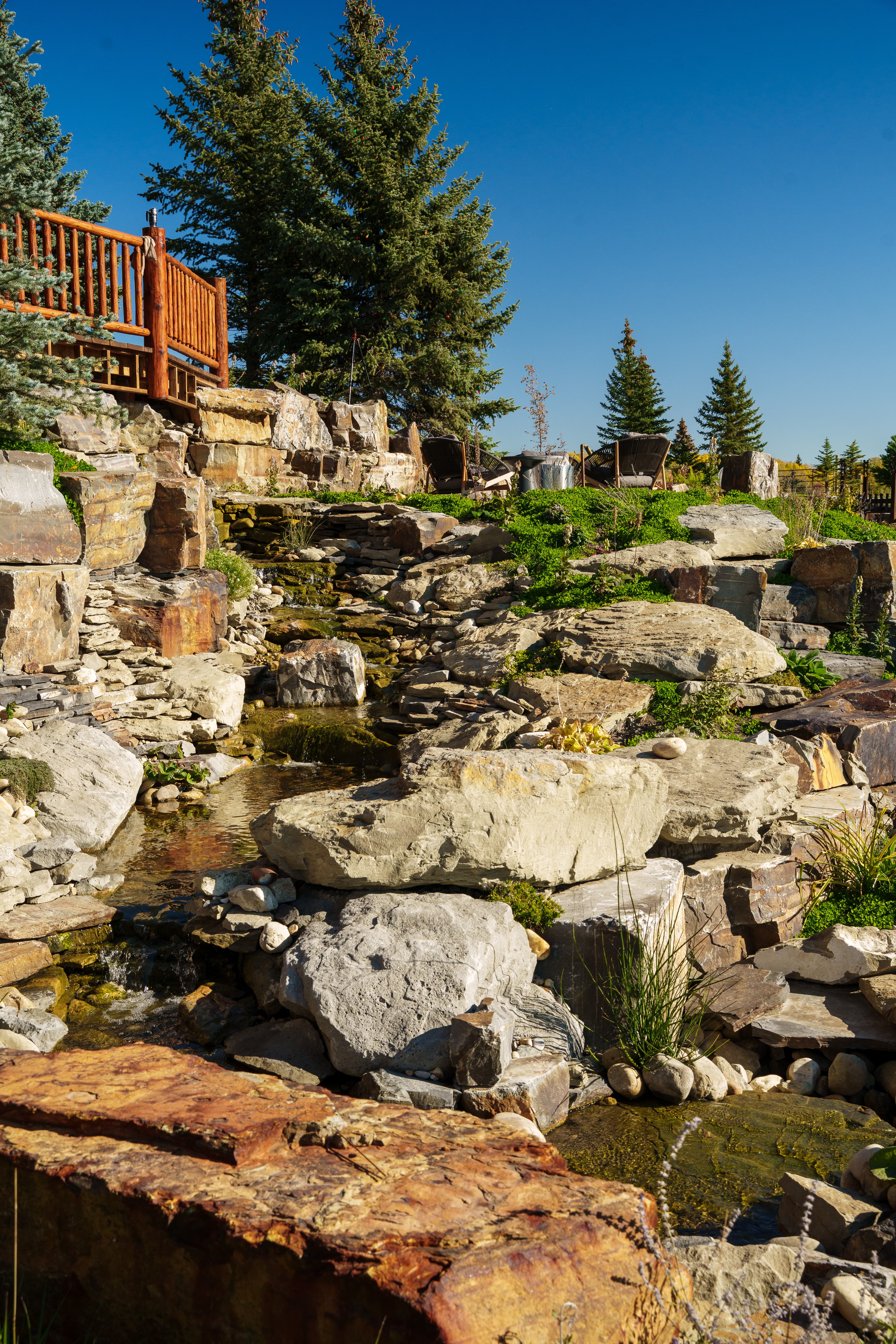Tiered stone waterfall in a lush garden with evergreen trees and a wooden deck.