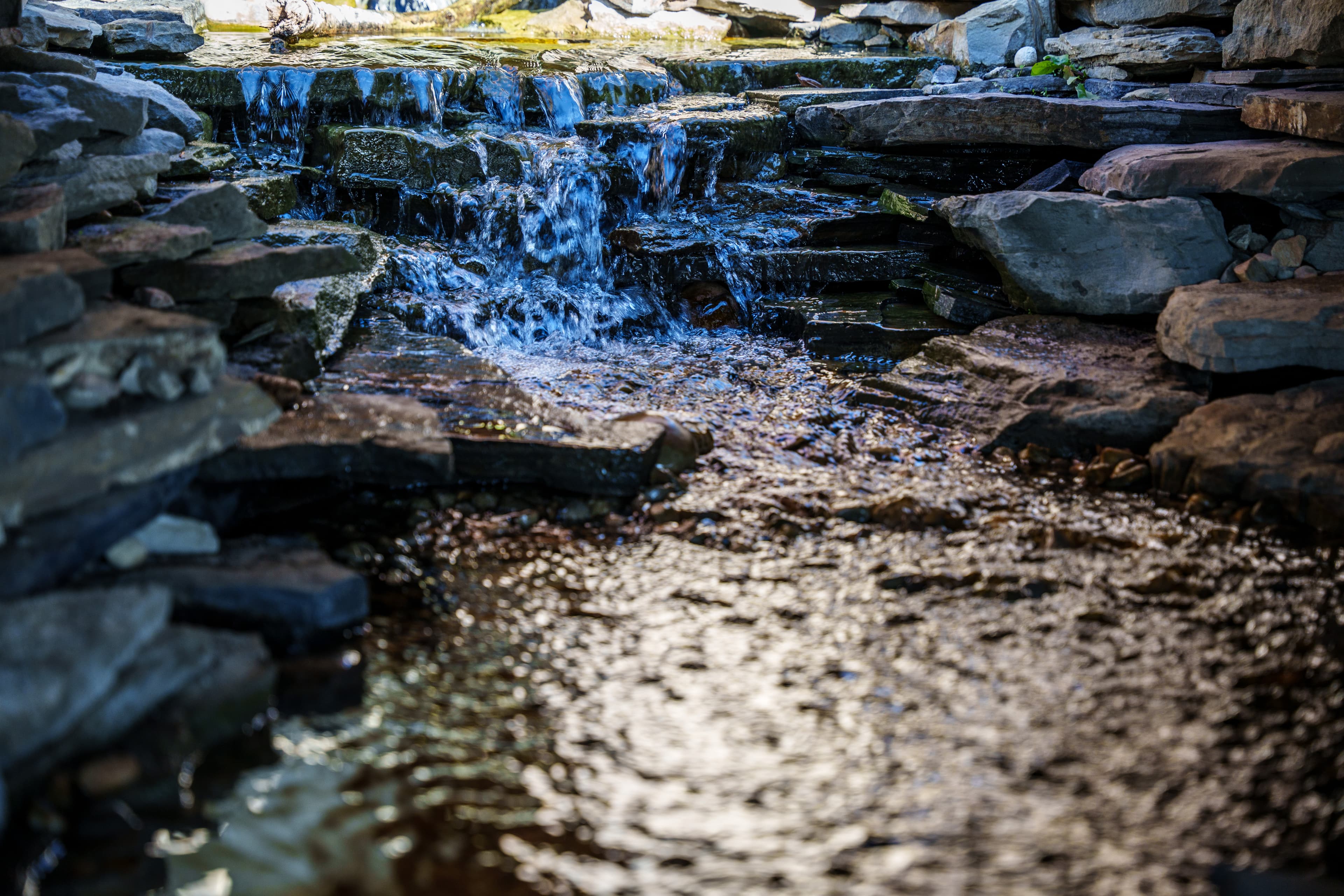 A small waterfall cascades over flat, layered stones into a shallow stream with sunlight reflections.
