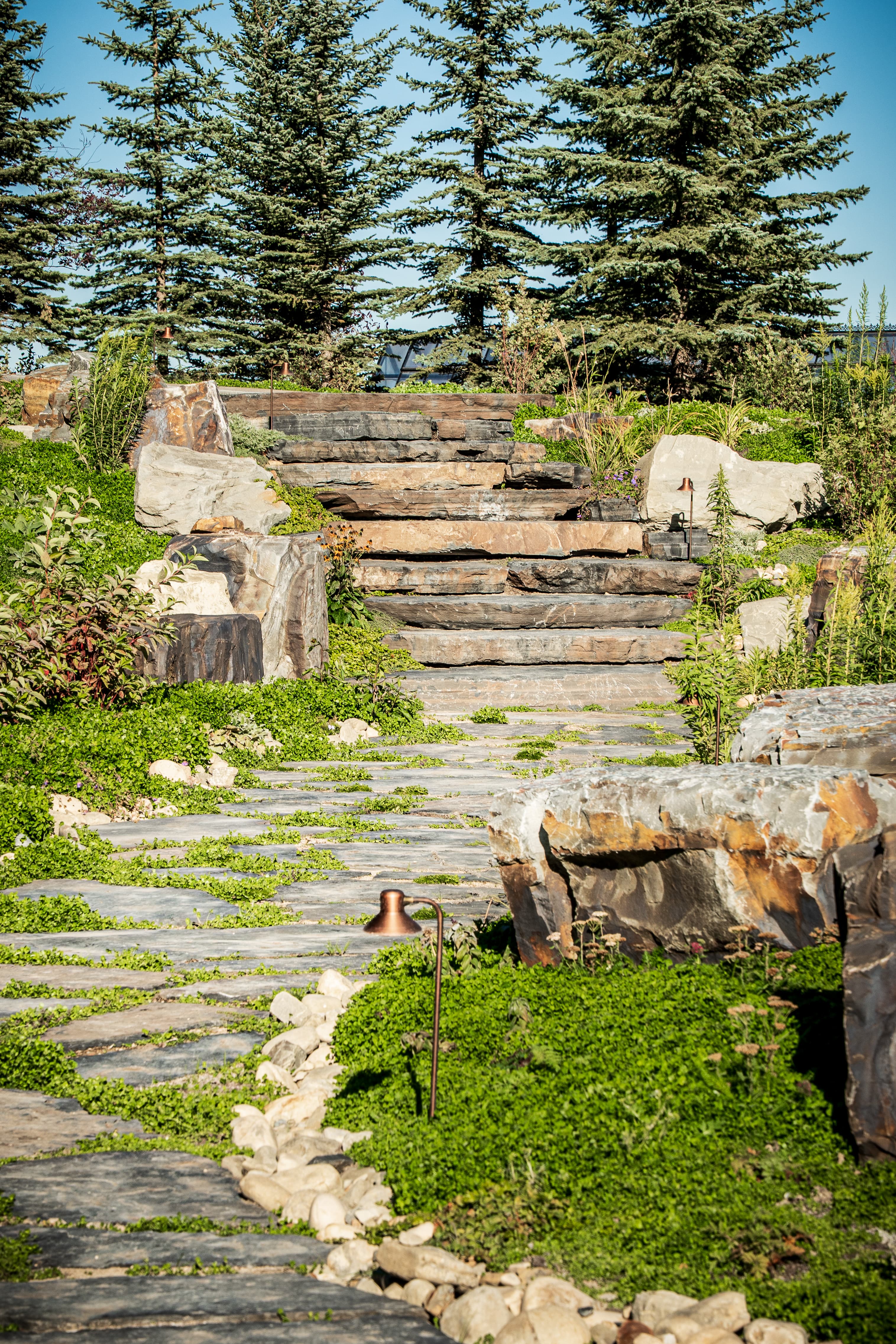 Stone path and steps lead through a lush garden with large rocks and tall evergreens.